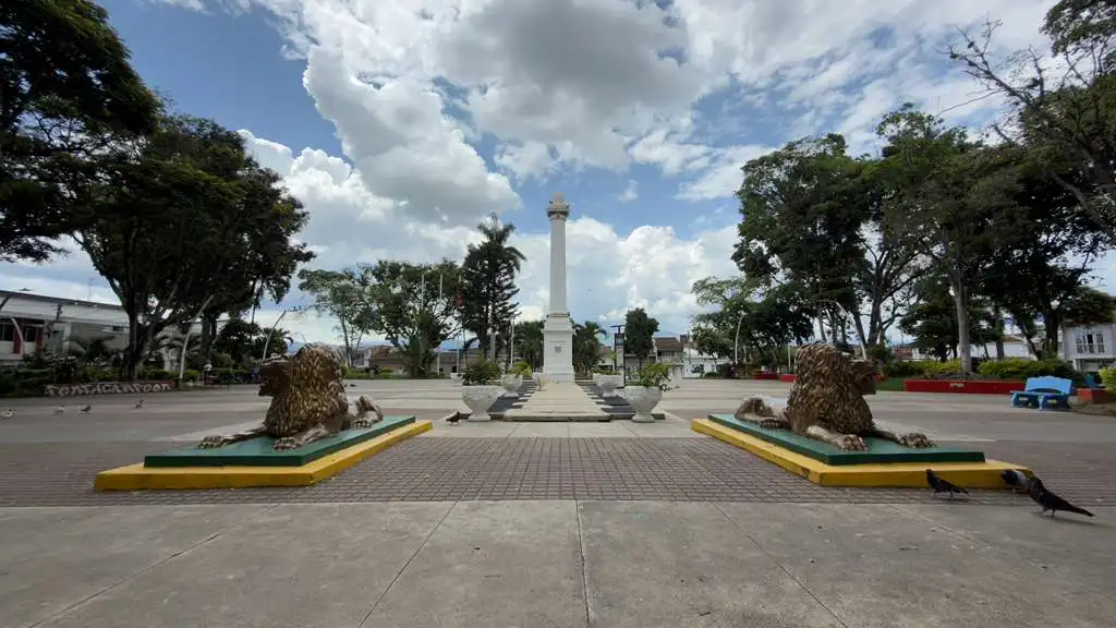 Parque principal de Buga con un monumento central y dos esculturas de leones, rodeado de árboles y edificios. Cielo nublado.
