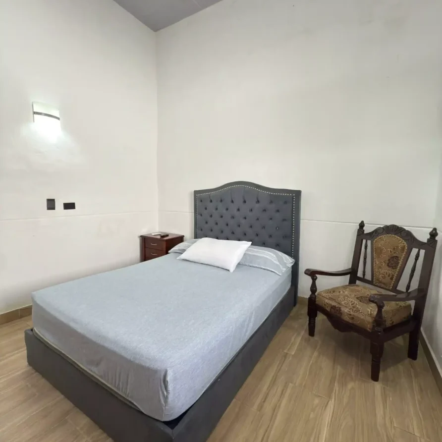 Habitación de hotel con cama doble, cabecero gris tapizado, mesita de noche y silla, cerca a la Basílica en Buga.